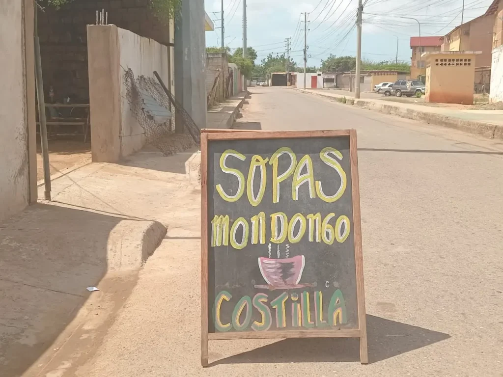 aviso de cartelera de pizarra con letras amaarillas y vees y se lee 2sopas mondongo costilla" en medio de la calle como imagen de fotorreportaje de avisos latinoamericanos de la seccion encuadre del medio digital el pajaro azul