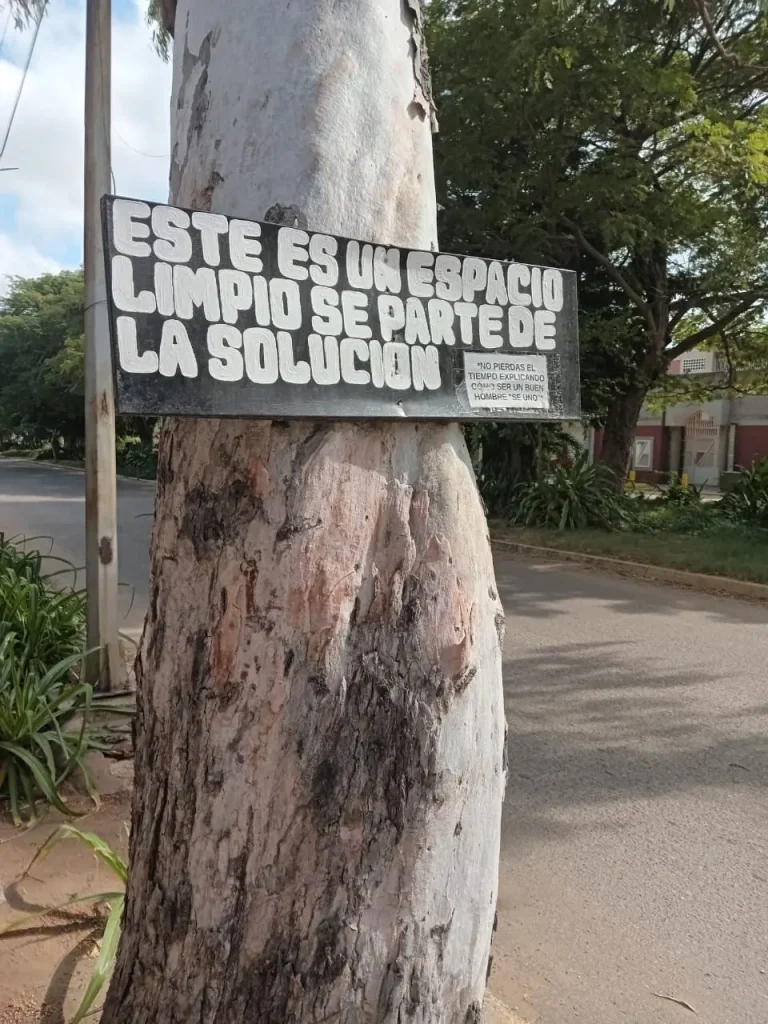Cartel callejero latinoamericano atado a un árbol con el mensaje “Este es un espacio limpio, sé parte de la solución”, fondo una calle con una plaza con arboles verdes fotografiado para el reportaje gráfico Encuadre de El Pájaro Azul